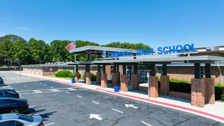 Front entrance of Campbell High School with brick pillars, American flag, and parked cars under clear blue sky.
