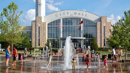 Children playing in water fountains in front of a large modern city hall building on a sunny day.