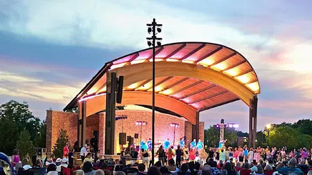 Outdoor concert at sunset under a modern wooden bandstand with a large audience gathered on lawn.
