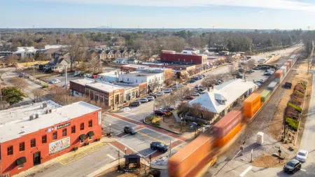 Aerial view of a city street with buildings, parked cars, and a train moving along the tracks in daylight.