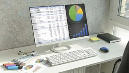 Modern workspace with computer displaying financial charts, keyboard, mouse, eyeglasses, markers, and notebooks on desk.