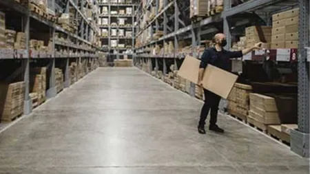 Worker wearing mask holding a box and organizing shelves in a large warehouse with stacked inventory.