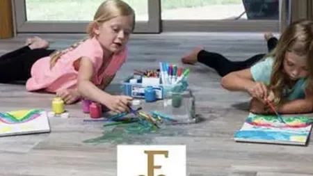 Two girls painting with watercolors on a wood-patterned floor inside a bright room with large windows