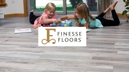 Two young girls painting and drawing on light wood flooring in a bright living room with large windows.