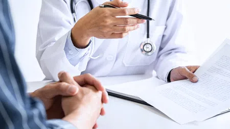 Doctor consulting patient, holding paper and pen, discussing medical information at a white desk