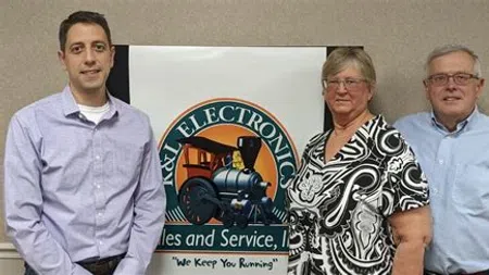 Three people standing beside a colorful I&I Electronics sign featuring a train logo indoors