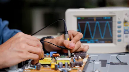 Hands testing an electronic circuit board using probes with an oscilloscope showing waveform in the background