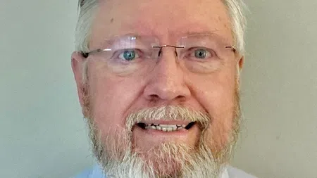 Smiling older man with a white beard and glasses wearing a light blue button-up shirt against a plain background.