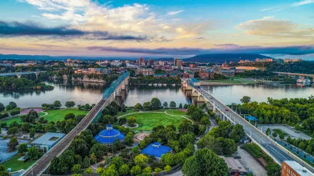 Aerial view of city with twin bridges over river, green park with blue-roofed buildings, and cloudy sky at sunset.
