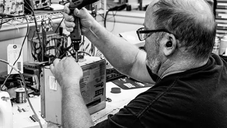 Man using a screwdriver to repair electronic equipment at a cluttered workbench in a workshop.