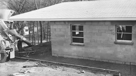 Construction site with a cinder block building, cement mixer truck pouring concrete, and workers at windows.
