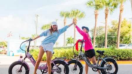 Two women high-fiving while standing next to electric bikes on a sunny street with palm trees.