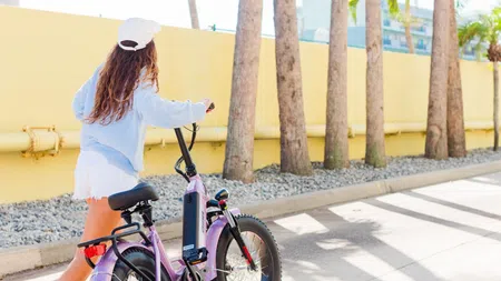 Woman in white cap and blue shirt walking a purple electric bike on sunny street with palm trees and yellow wall.