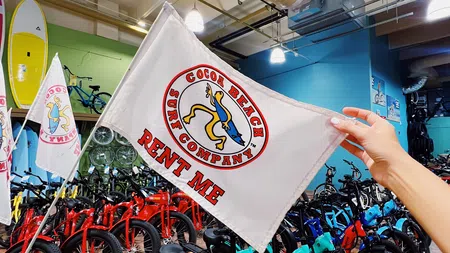 Hand holding a Cocoa Beach Surf Company flag in front of colorful rental bikes inside a shop with surfboards.