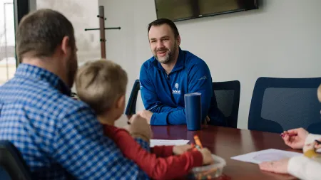 Man in blue jacket smiling and talking to child and adult seated at a conference table during meeting.