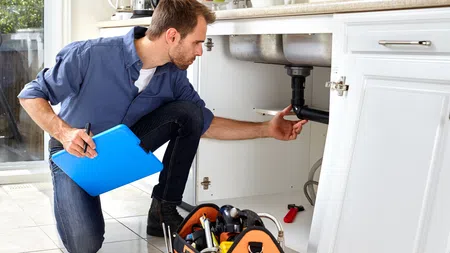 Plumber inspecting kitchen sink pipes with tools and clipboard, performing maintenance or repair work.