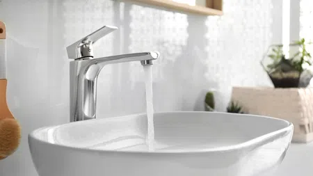 Modern chrome faucet pouring water into a sleek white bathroom sink with decorative plants in the background