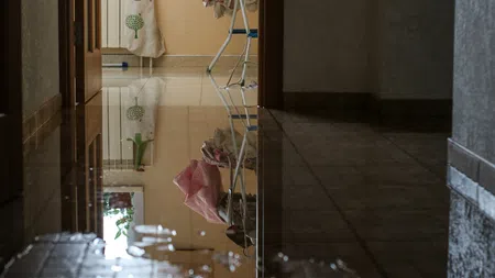 Indoor hallway flooded with water reflecting furniture, clothes, and a window with plant outside, highlighting water damage.