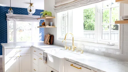 Bright modern kitchen with white cabinets, marble countertops, gold fixtures, and large windows with natural light.