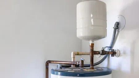 Residential water heater with expansion tank and copper piping installed on white wall in utility room.