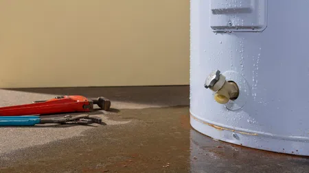 Leaking water heater with rusty base and plumbing tools on wet concrete floor indoors.