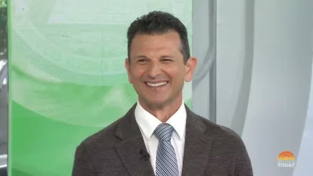Smiling man in a brown suit and patterned tie speaking on a green and white news set
