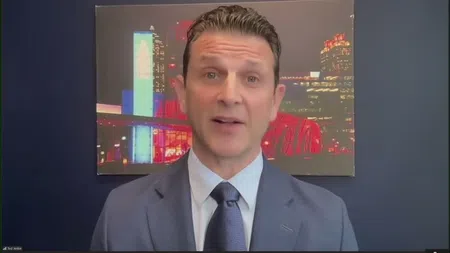 Man in navy suit and tie speaking with cityscape artwork behind him against dark blue wall