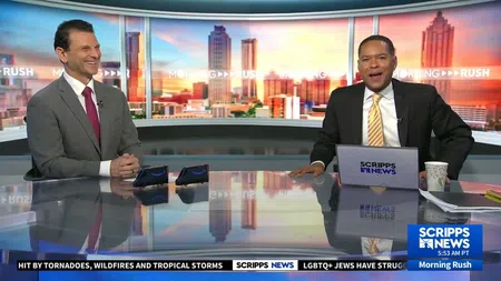 Two male news anchors in suits hosting Scripps News Morning Rush with city skyline background at sunrise.