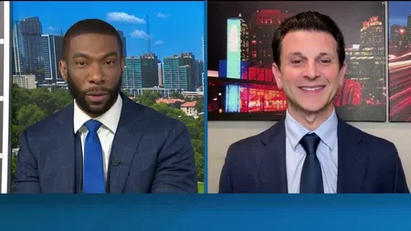 Two male news anchors in suits conducting a split-screen interview with city skylines in the background.