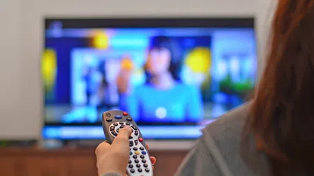 Person holding a TV remote control pointing at a television screen showing a blurred image of a woman.