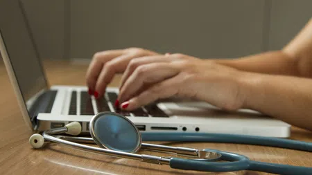 Hands typing on a laptop with a blue stethoscope resting on the wooden desk in a medical workspace.