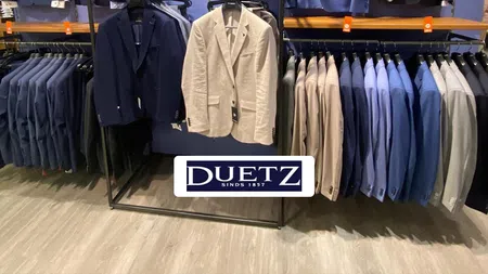 Men's suits and blazers in various colors displayed on racks in a Duetz clothing store with a blue wall background.