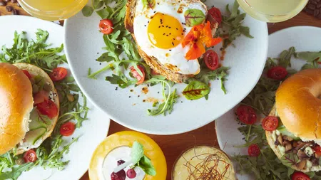 Brunch spread with egg-topped toast, veggie burgers, fresh greens, cherry tomatoes, and colorful beverages on wooden table.