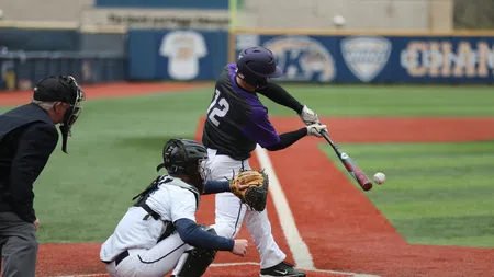 Baseball batter in purple jersey swings at pitch with catcher and umpire behind home plate on field.