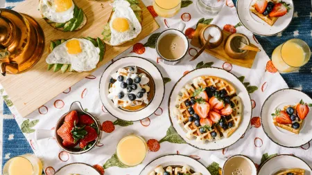 Breakfast spread with waffles topped with strawberries and blueberries, avocado toast with eggs, orange juice, and coffee.