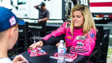 Female race car driver in pink suit signing autographs for a young fan at an IndyCar event.