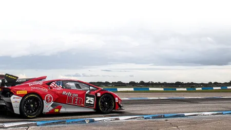 Red and white race car number 25 speeding on a wet racetrack under cloudy skies.