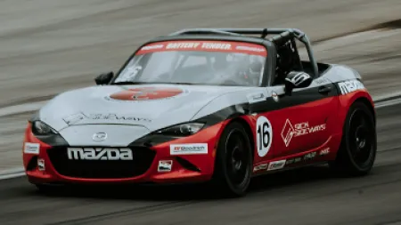 Red and white Mazda race car number 16 speeding on a race track with sponsor logos and black wheels.