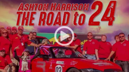 Group of people in red shirts with a red race car and banner reading Ashton Harrison The Road to 24