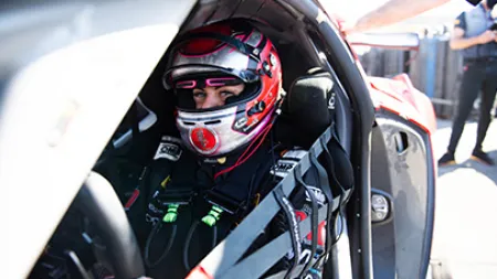 Race car driver in red helmet sitting inside a racing car with safety harness fastened.