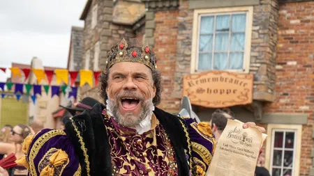 Man dressed as a medieval king entertains two young girls at a Renaissance fair with a mug and parchment.