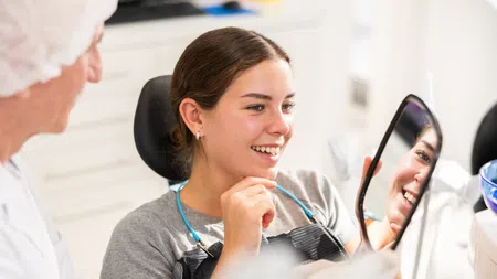 Young woman smiling while looking at her teeth in a hand mirror at the dentist office with the dentist nearby.