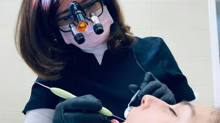 Dentist wearing magnifying glasses and gloves treating patient lying in dental chair with tools.