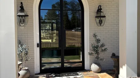 Front entry with black arched glass door, stone steps, white brick walls, potted plants, and wall lanterns