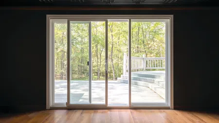 Sliding glass doors open to a wooden deck with white railing and a forest view outside during daytime