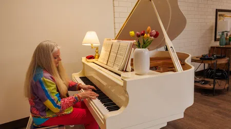 Elderly woman playing a white grand piano in a cozy room with music sheets and a vase of tulips.
