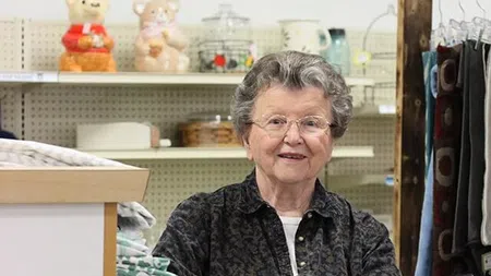 Senior woman shopping at a thrift store, holding fabric with shelves of items in the background.