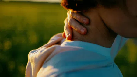Person in soft light holding neck with hand, outdoors with blurred green background at sunset.