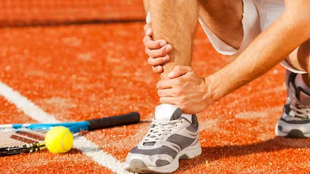 Tennis player holding injured ankle on clay court near racket and ball by the white line.