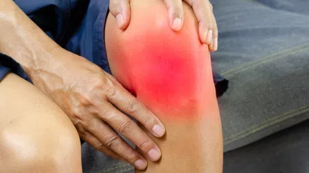 Person holding their knee with visible redness indicating pain or inflammation while sitting on a couch.